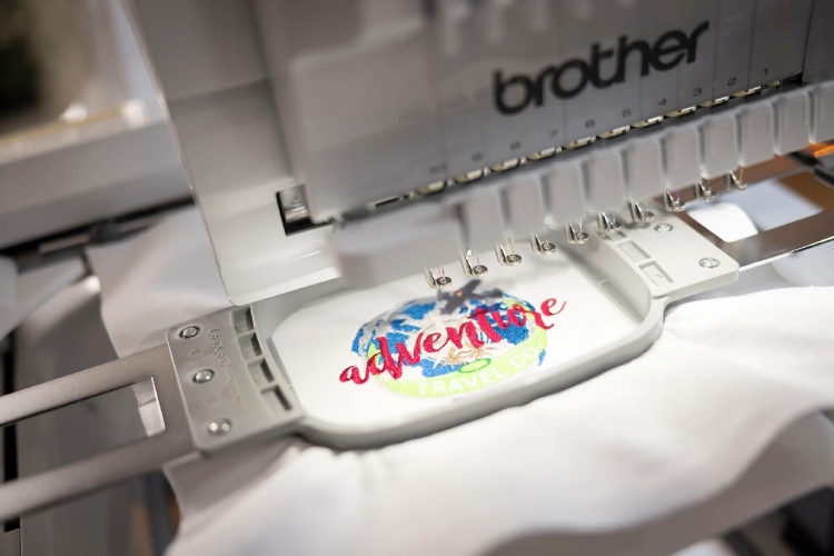 Program Brother BE1 EmbroideryStudio do projektowania haftów, z bliska na maszynę do haftu, z widocznym haftem adventure w kolorze czerwonym oraz grafiką ziemi w odcieniach niebieskiego i zielonego, na białym tle materiału.
