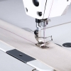 Jednoigłowa stebnówka TEXI TRONIC 1F PREMIUM EX, z bliska, szyjąca przez beżowy materiał. Widoczna stopka i igła, z różowym nitkiem prowadzącym przez materiał, z szarym elementem na stole maszyny.
