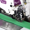 Owerlok TEXI QUATTRO 3F PREMIUM EX w zbliżeniu, z widocznym stopek i igłą, szyjący zielony materiał. Na maszynie logo texi oraz elementy sterujące. Stół maszyny z metaliczną powierzchnią.