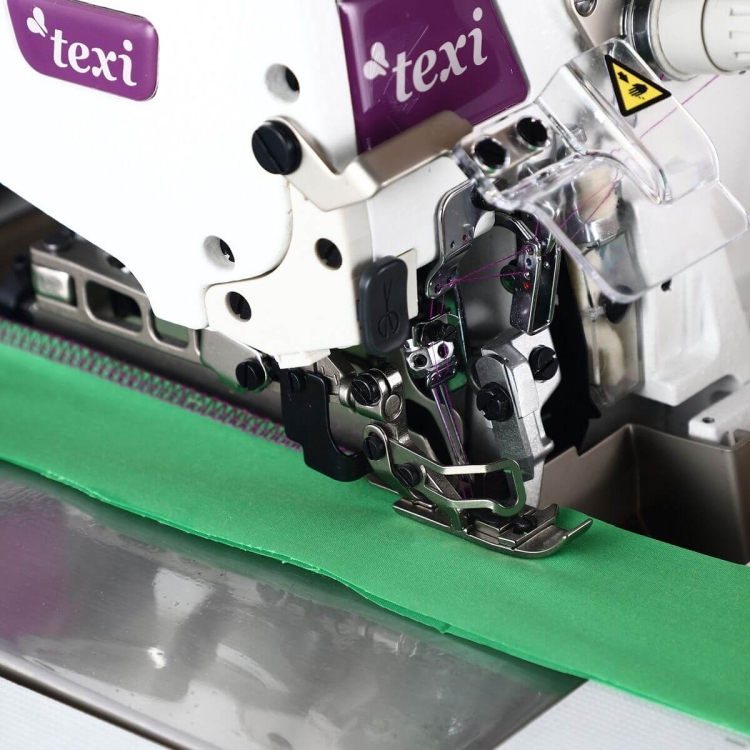 Owerlok TEXI QUATTRO 3F PREMIUM EX w zbliżeniu, z widocznym stopek i igłą, szyjący zielony materiał. Na maszynie logo texi oraz elementy sterujące. Stół maszyny z metaliczną powierzchnią.
