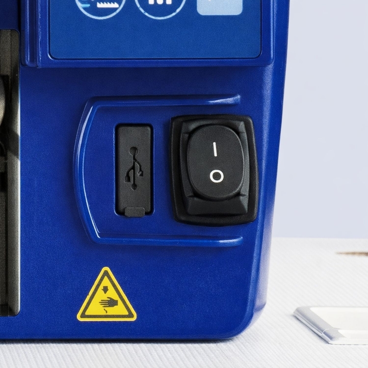Przycisk zasilania oraz port USB na obudowie stebnówki JUKI DDL-7000AS, w kolorze niebieskim. Widoczna również żółta etykieta ostrzegawcza z symbolem ręki. Ujęcie frontalne, akcentujące detale panelu sterującego.