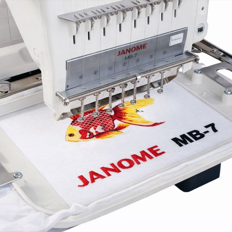 Na zdjęciu ukazana jest igielnica hafciarki Janome MB-7E. Na tamborku jest założony materiał który jest haftowany. Hafciarka wyhaftowała już na materiale kolorową rybę wraz z napisem JANOME MB-7E.
