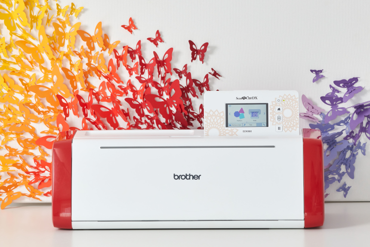Ploter tnący Brother ScanNCut SDX900 w białej obudowie z czerwonym akcentem, z wyświetlaczem dotykowym, umieszczony na tle kolorowych, wyciętych motyli w odcieniach żółci i czerwieni.
