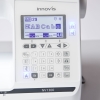 Zbliżenie na cyfrowy ekran LCD maszyny do szycia Brother NV1300, który wyświetla wzory liter i parametry ściegu. Poniżej znajduje się panel z przyciskami numerycznymi i funkcyjnymi, a obok podświetlane ikony pozycjonowania igły i obcinania nitki.