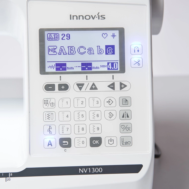 Zbliżenie na cyfrowy ekran LCD maszyny do szycia Brother NV1300, który wyświetla wzory liter i parametry ściegu. Poniżej znajduje się panel z przyciskami numerycznymi i funkcyjnymi, a obok podświetlane ikony pozycjonowania igły i obcinania nitki.