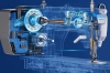 Zdjęcie pokazuje techniczny przekrój maszyny z mocno wyeksponowanym silnikiem, wałem napędowym, elektroniką i mechanizmem igielnicy. Grafika podkreśla zintegrowanie napędu z głowicą oraz elektroniczne sterowanie pracą maszyny.