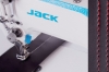 Przemysłowa stebnówka JACK H5K podczas wykonywania równoległych przeszyć na czarnym materiale. Po prawej stronie pokazano zbliżenie równych szwów wykonanych kontrastową czerwoną nicią na powierzchni materiału.