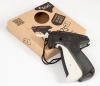 Pistolet do etykietowania Texi 75F GUN fine w kolorze czarno-białym, z widoczną rękojeścią i przyciskiem. Obok znajduje się opakowanie kartonowe z okrągłym otworem oraz grafiką produktu. Produkt leży na białym tle.