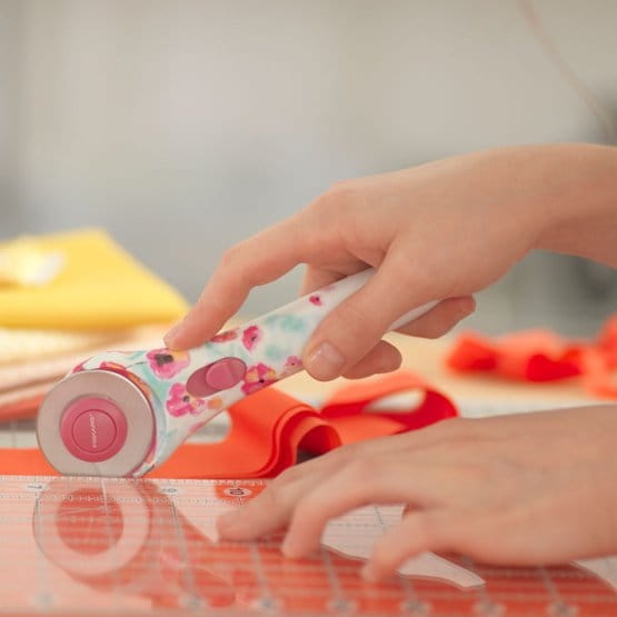 Nóż krążkowy Fiskars Inspiration Flower 1023907 o średnicy 45 mm, z kwiatowym wzorem na uchwycie, trzymany w dłoni, na przezroczystej linijce. W tle widoczne kolorowe materiały krawieckie, w tym żółty i czerwony.