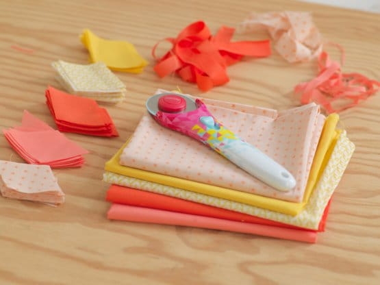 Nóż krążkowy Fiskars Inspiration Geometric 1023908 z kolorowym uchwytem leży na stosie tkanin w odcieniach pomarańczowym i żółtym. W tle widoczne kawałki materiału oraz wstążki, na drewnianym blacie.