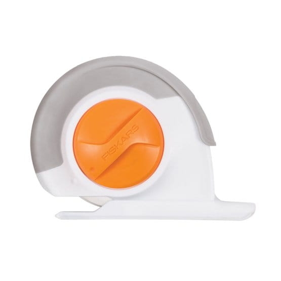 Nóż krążkowy krawiecki Fiskars Chenille o średnicy 60 mm, widoczny z boku. Posiada ergonomiczną rączkę w szaro-białej kolorystyce oraz pomarańczowy przycisk blokady. Stojący na płaskiej powierzchni, idealny do precyzyjnego cięcia materiałów.