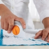 Nóż krążkowy krawiecki Fiskars Chenille o średnicy 60 mm, trzymany w dłoni, używany do cięcia materiału. Narzędzie ma szary uchwyt z pomarańczowym przyciskiem, a tło to niebieska tkanina. Kąt ujęcia: z boku, z bliska, skupiony na detalu użytkowania.