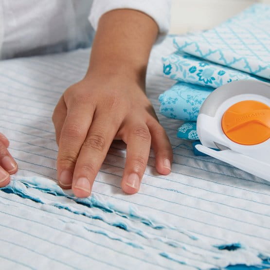 Nóż krążkowy krawiecki Fiskars Chenille 1023906 o średnicy 60 mm leży obok złożonych tkanin w błękitne wzory. Na pierwszym planie widoczna jest dłoń użytkownika trzymająca tkaninę na stole, co sugeruje użycie narzędzia do cięcia materiału.