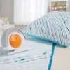 Nóż krążkowy krawiecki Fiskars Chenille 1023906 o średnicy 60 mm, w kolorze białym z pomarańczowymi akcentami, leży na niebieskiej tkaninie z widocznymi przeszyciami. W tle zmięta poduszka. Produkt pokazany w aranżacji krawieckiej, na jasnym tle.