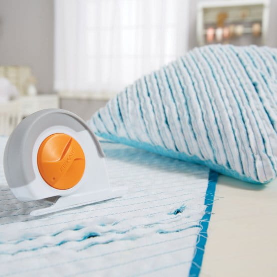 Nóż krążkowy krawiecki Fiskars Chenille 1023906 o średnicy 60 mm, w kolorze białym z pomarańczowymi akcentami, leży na niebieskiej tkaninie z widocznymi przeszyciami. W tle zmięta poduszka. Produkt pokazany w aranżacji krawieckiej, na jasnym tle.