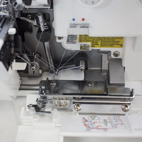 Część wewnętrzna owerloka JUKI MO-644D z widocznymi nitkami i mechanizmem szyjącym. Ujęcie z boku ukazuje metalowe elementy, dźwignie oraz instrukcje na obudowie. Złożony w mechanizmie stoper oraz prowadnik nitki.