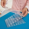 Składana linijka Fiskars 1023902 do patchworku i quiltingu, przezroczysta, z nadrukowanymi liniami i cyframi, trzymana w dłoni. Ułożona na niebieskim tle, pokazuje dwie warstwy z widocznymi oznaczeniami.