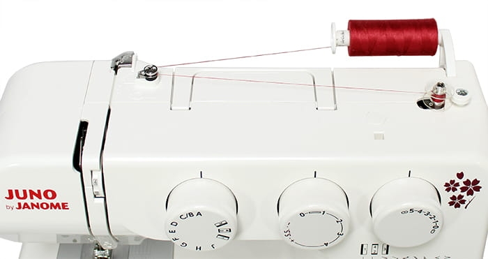 Maszyna do szycia Janome JUNO E1019 w kolorze białym z widoczną czerwoną szpulą. Posiada trzy pokrętła do regulacji, logo JUNO by JANOME oraz detale ułatwiające nawlekanie nici. Ujęcie z góry ukazujące górną część maszyny.