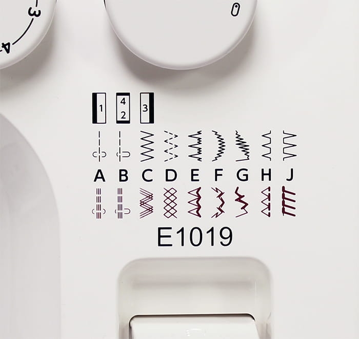 Panel maszyny do szycia Janome JUNO E1019, z widocznymi pokrętłami do ustawiania ściegów oraz schematem dostępnych wzorów ściegów oznaczonych literami A-J i numerami 1-3. Kolorystyka dominująca: biała z czarnymi i bordowymi detalami.