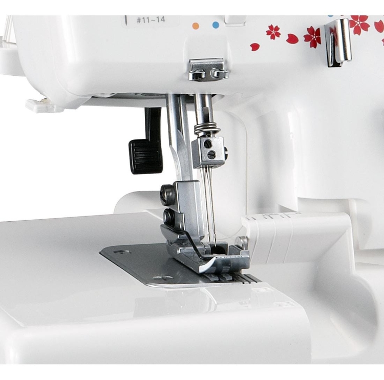 Owerlok Janome 990D z widoczną stopką i igłą w pozycji roboczej. Maszyna w kolorze białym z dekoracyjnymi kwiatami. Zbliżenie na mechanizm szyjący, z uchwytem do regulacji oraz widocznymi oznaczeniami na obudowie.