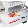 Hafciarka Janome MB-7E z 7 igłami, ukazana z przodu, haftująca kolorowy wzór złotej rybki na białym materiale. W dolnej części widoczny napis JANOME MB-7 w czerwonym kolorze. Maszyna na białym tle, z widocznymi przewodami i mechanizmem haftującym.