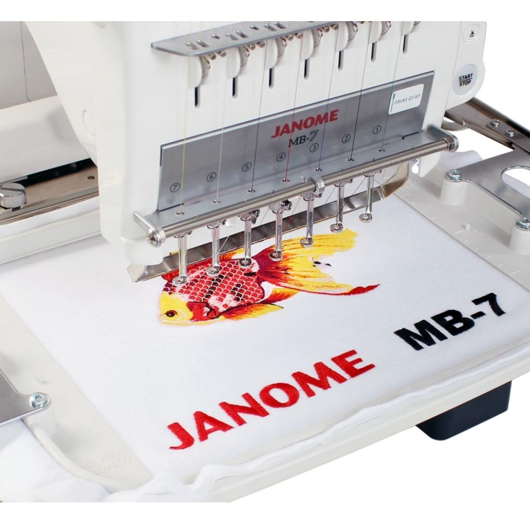 Hafciarka Janome MB-7E z 7 igłami, ukazana z przodu, haftująca kolorowy wzór złotej rybki na białym materiale. W dolnej części widoczny napis JANOME MB-7 w czerwonym kolorze. Maszyna na białym tle, z widocznymi przewodami i mechanizmem haftującym.