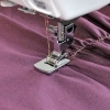 Stopka do maszyny Janome 7, chwytacz rotacyjny 9mm, w zbliżeniu na metalową stopkę z widocznym nicią, używaną do szycia na fioletowym materiale. Produkt umieszczony w pozycji pionowej, z bliska uwidacznia detale konstrukcyjne.