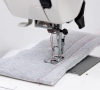 Maszyna do szycia Janome HD 523 EASY JEANS z bliska, z widoczną stopką do szycia na grubszych materiałach. Na szarej tkaninie widoczna jest czerwona nitka. Ujęcie pokazuje igłę, mechanizm i płytę maszyny na białym tle.