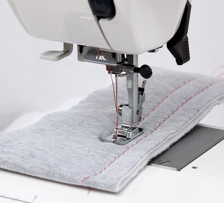 Maszyna do szycia Janome HD 523 EASY JEANS z bliska, z widoczną stopką do szycia na grubszych materiałach. Na szarej tkaninie widoczna jest czerwona nitka. Ujęcie pokazuje igłę, mechanizm i płytę maszyny na białym tle.
