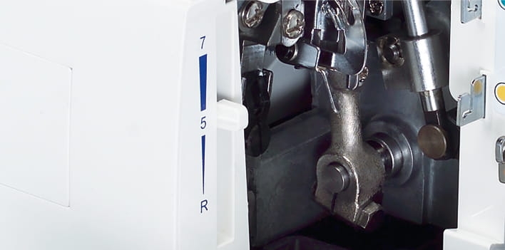 Zbliżenie na mechanizm owerloka Minerva M840DS, ukazujące metalowe elementy, pokrętło z oznaczeniami 5 i 7 oraz przyciski funkcyjne. Widoczny jasny korpus maszyny w kolorze białym, z częściami roboczymi w środku.