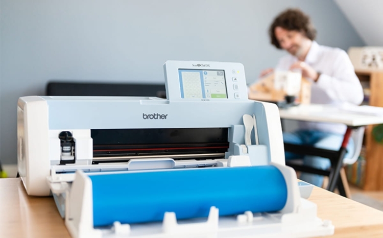 Podajnik do cięcia folii Brother ScanNcut CADXRF2 w kolorze niebieskim, z widoczną jednostką tnącą i ekranem dotykowym. Ujęcie frontalne, produkt umieszczony na stole, w tle pracujący mężczyzna.