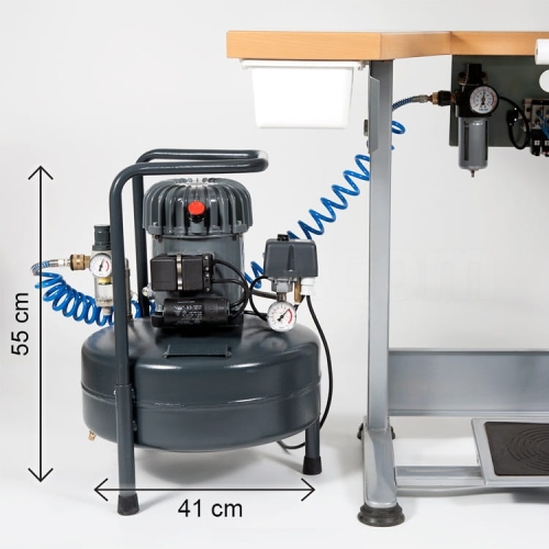 Sprężarka SIL-EOL 24/50 o pojemności 24l, wysokości 55 cm i średnicy 41 cm, z tłokowym mechanizmem. W kolorze ciemnoszarym, z manometrem i niebieskim wężem powietrznym, umieszczona obok stołu roboczego. Maksymalne ciśnienie 8 bar, wydajność 50 L/min.