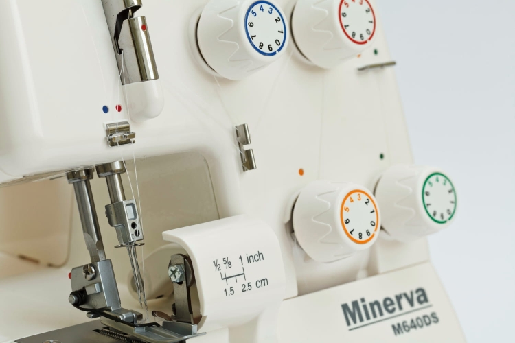 Owerlok Minerva M640DS 4-nitkowy, z widocznymi białymi pokrętłami regulacyjnymi w kolorach: niebieskim, pomarańczowym i zielonym. Na obudowie umieszczone są oznaczenia pomiarowe. Maszyna stoi w pozycji otwartej, z widoczną igłą i mechanizmem szyjącym.