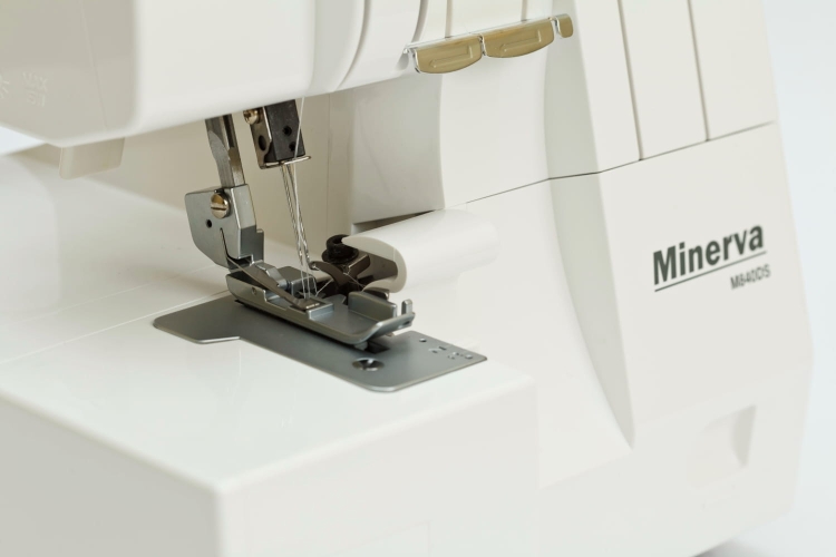 Owerlok Minerva M640DS 4-nitkowy z widokiem na stopkę i igłę. Maszyna ma białą obudowę i metalową płytkę pod stopką. Widać mechanizm naciągu nici oraz logo Minerva na obudowie. Ujęcie ze zbliżenia, ukazujące detale pracy maszyny.