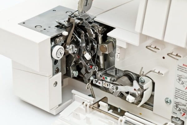 Zbliżenie na wnętrze overlocka Coverlok Minerva M5000CL, ukazujące mechanizm szyjący z widocznymi igłami, napinaczami i elementami regulacyjnymi. Obudowa maszyny jest biała, metalowe detale kontrastują z plastikiem.