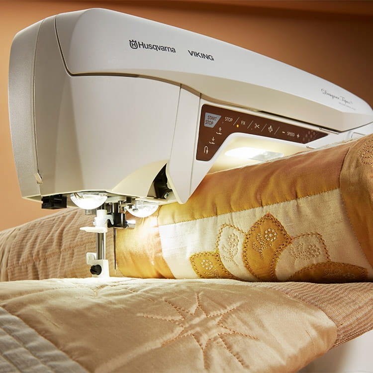 Maszyno-hafciarka Husqvarna Designer Topaz 25 w kolorze białym, z widocznymi przyciskami sterującymi i oświetleniem. Produkt umieszczony na tkaninie z haftem w odcieniach złota i beżu, w zbliżeniu na detal szycia.