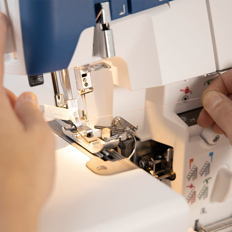Owerlok Husqvarna AMBER AIR S400 z automatycznym systemem nawlekania, z widoczną igłą oraz mechanizmem nawlekania. Ujęcie z boku, z ręką trzymającą nitkę. Na obudowie umieszczone symbole ułatwiające obsługę. Kolor biały z niebieskimi akcentami.