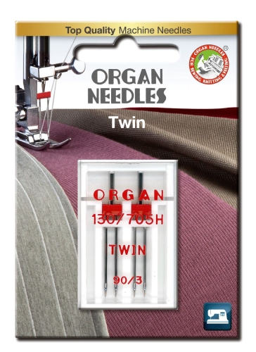 Igła podwójna półpłaska Organ TWIN 130/705H 90/3mm w blistrze, zawierająca dwie igły. Opakowanie z przezroczystego plastiku, widoczne czerwone napisy ORGAN oraz TWIN. Tło w odcieniach szaro-fioletowych, z maszyną do szycia w górnej części zdjęcia.