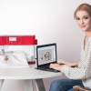 Maszyno-hafciarka PFAFF CREATIVE 1.5 K100 w kolorze czerwonym i białym, z górnym transportem, umieszczona na stole obok laptopa, na którym widoczny jest projekt haftu. Użytkownik w bluzce w kropki pracuje przy maszynie.