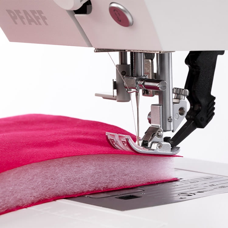 Maszyna do szycia PFAFF QUILT AMBITION 630, widok z bliska na stopkę i igłę, szyjąca różowy materiał z ociepliną. Widoczny podwójny transport i panel sterowania. Tło białe.