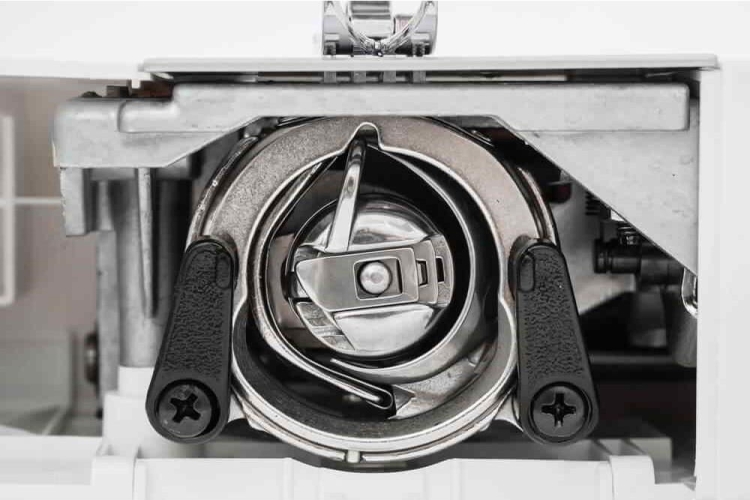 Zbliżenie na chwytacz maszyny do szycia Minerva M832B. Widoczny metalowy chwytacz z plastikowymi uchwytami po bokach. Elementy mocujące i mechanizmy wewnętrzne, z jasnym tłem.