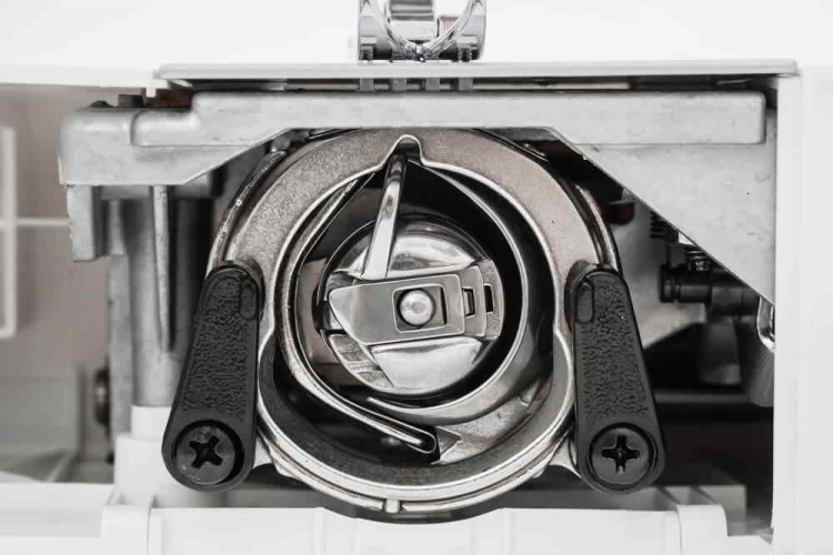 Widok z bliska na mechanizm bębenka maszyny do szycia Minerva M819B. Metalowy bębenek z plastikowymi uchwytami po bokach, widoczny element blokujący. Całość w jasnej obudowie, z detalami chromowanymi, ukazująca precyzyjne wykonanie.