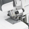Zbliżenie na mechanizm owerloka TEXI TEGRA 4, pokazujące cztery nici, szare metalowe elementy, oraz białą szpulę. Widoczne są detale stopy oraz obszar roboczy maszyny. Tło jest neutralne, co podkreśla produkt.
