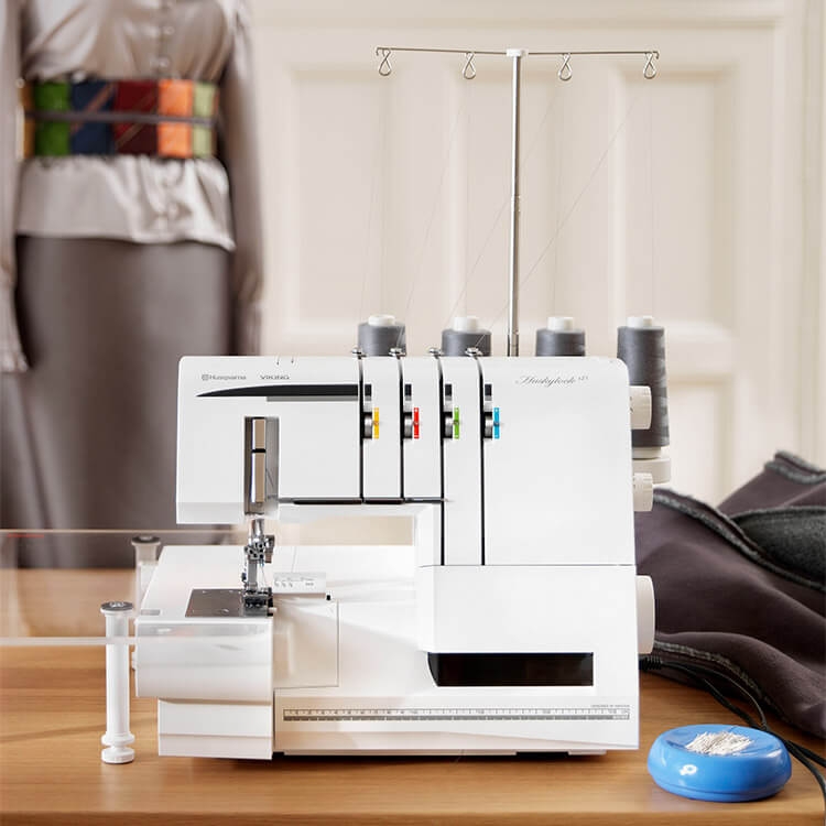 Overlock Husqvarna Viking HUSKYLOCK S21 w kolorze białym z funkcją renderki. Urządzenie z czterema szpulkami, widocznym stół roboczym oraz regulatorem naprężenia nici. W tle manekin z odzieżą oraz akcesoria krawieckie, na drewnianym blacie.