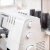 Overlock Husqvarna Viking HUSKYLOCK S21 w kolorze białym z widocznymi pokrętłami do regulacji. Zestaw kolorowych szpulek w tle. Maszyna stoi na stole, w bliskim ujęciu z boku, ukazując detale konstrukcji.