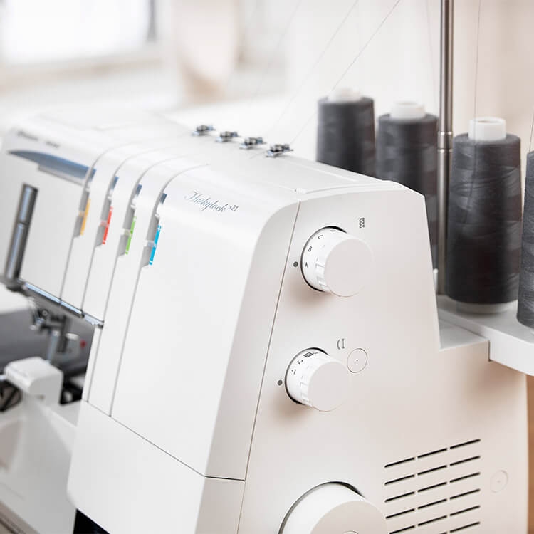 Overlock Husqvarna Viking HUSKYLOCK S21 w kolorze białym z widocznymi pokrętłami do regulacji. Zestaw kolorowych szpulek w tle. Maszyna stoi na stole, w bliskim ujęciu z boku, ukazując detale konstrukcji.