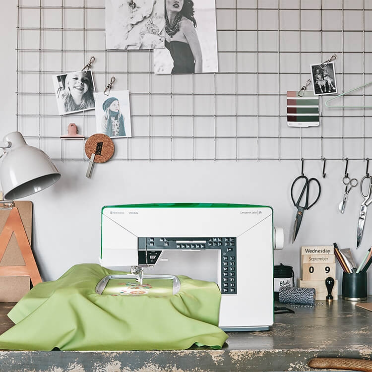 Maszyno-hafciarka Husqvarna Viking DESIGNER JADE 35 w kolorze białym z zielonymi akcentami, z widocznym haftem na zielonym materiale. Ujęcie z boku, na stole z dodatkami jak lampka, nożyczki i zdjęcia na tle siatki.