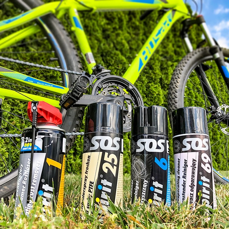 Zestaw do konserwacji roweru Spirit składa się z czterech sprayów: odtłuszczacza, smaru PTFE, środka czyszczącego i preparatu do ochrony. Butelki stoją na trawie, w tle widoczny jest zielony rower górski.