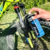 Zestaw do konserwacji roweru Spirit, zawierający cztery spray''e, używany do czyszczenia amortyzatora. Widoczna ręka trzymająca spray z niebieską etykietą, na tle zielonej trawy i części roweru. Amortyzator pokryty pianą czyszczącą.