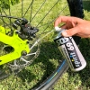 Zestaw do konserwacji roweru, w tym spray Spirit 69, trzymany w dłoni, aplikowany na hamulec hydrauliczny. W tle widoczna opona rowerowa i trawa. Spray w metalowej puszce z wyraźnym napisem. Kolor puszki: srebrny z czarnymi i kolorowymi detalami.